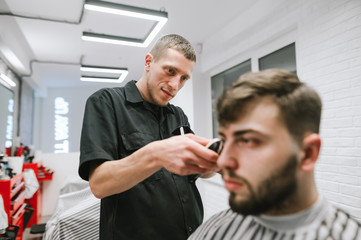 Professional barber cuts a bearded client in a light barber shop, looks at her hair and uses a clipper. Positive male hairdresser makes male haircut with a clipper. Barbershop concept