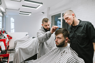 Two barbers are cutting a bearded man in a light modern barber shop. Hairdresser teacher teaches student to do hairstyles. Barber Workshop. Barber shop concept.