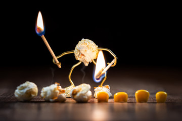 funny popcorn figure is holding a burning match on some corns to pop them up - obrazy, fototapety, plakaty