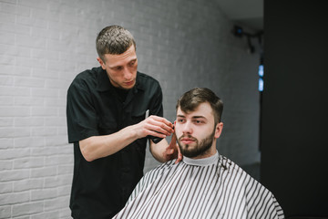 Handsome man with beard cuts hair in professional barber. Male hairdresser with serious face cuts client's hair clipper. Close up portrait of client and hairdresser at work.