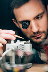 Selective focus of handsome clockmaker working with wristwatch on movement holder isolated on black