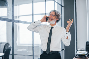 Mature businessman with grey hair and beard in formal clothes have conversation by the phone in the office