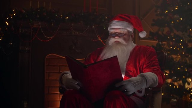 Senior man in santa clause clothing sitting in atmospheric room, reading a magical shining book with red cover - christmas spirit, holidays and celebrations concept.