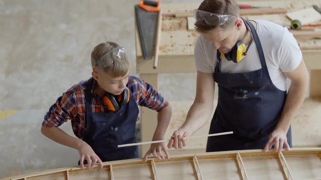 From above tilt up of father and son building wooden boat together standing at workbench in carpentry shop. Carpenter teaching kid how to attach strip of wood to form boat