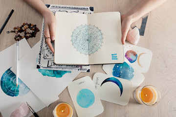 Top view of astrologer holding notebook with watercolor drawings and zodiac signs on cards on table - obrazy, fototapety, plakaty