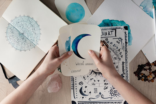 Top view of astrologer holding watercolor paintings with moon phases on cards by birth chart on table