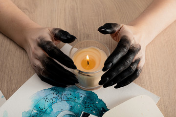 Cropped view of witch with black paint on hands holding candle by cards with watercolor zodiac...