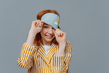Playful woman with ginger hair looks from sleep mask, wears striped pajamas, has cheerful expression, poses over grey background, enjoys sleeping at home. Healthy sleep and relaxation concept
