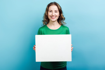 Blonde girl wearing in green sweater holding a white board copy space against blue background