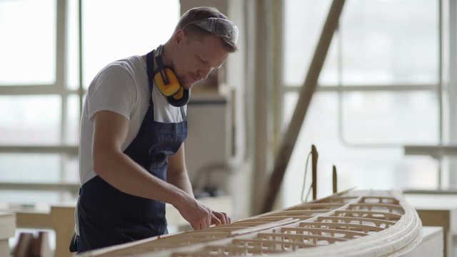 Side view medium shot of skilled middle aged carpenter making large wooden model of boat standing at workbench in woodworking shop
