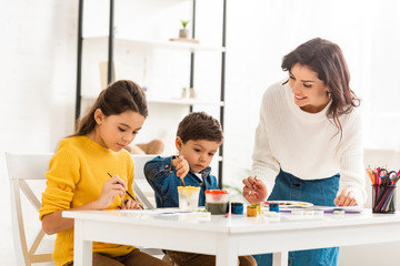happy woman standing near table and drawing with paints together with children