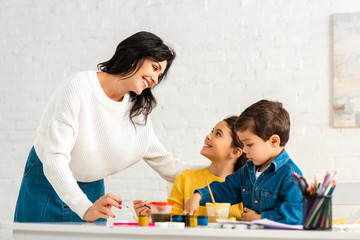 happy mother looking at children sitting at table and drawing with paints