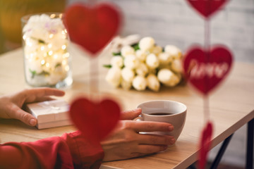 Valentines day concept. Cropped of lonely woman sitting on the table drinking coffee with bouquet of flowers tulips celebrating in restaurant. View through a heart-shaped garland.