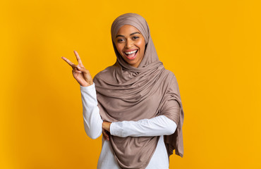 Joyful black muslim girl in headscarf showing peace gesture - obrazy, fototapety, plakaty