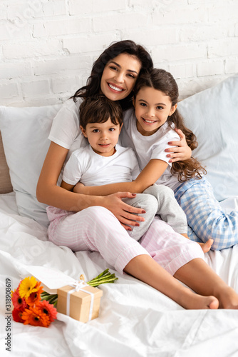 happy mother hugging adorable children while sitting in bed near bouquet and mothers day gift