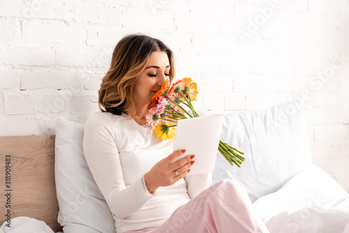 happy woman holding bouquet of gerberas and mothers day card while sitting in bed