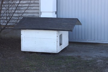 one white empty doghouse made of wooden boards and a gray roof stands on the ground and grass against the metal wall of the fence on the street