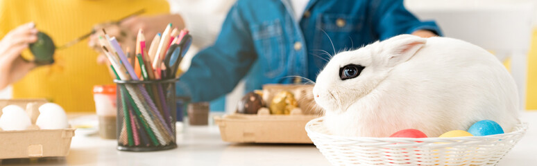 cropped view of brother and sister sitting at table with Easter eggs and white rabbit in wicker
