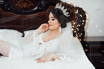 Beautiful model with bridal makeup and hairstyle. Morning of the bride, fees. Young beautiful girl lies on bed in white robe and underwear on the background of luxurious bedroom.
