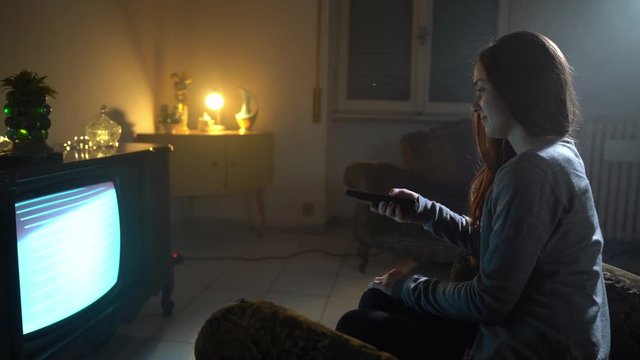 Abandoned dusk room with pile of antiques and smiling girl watching retro TV. Mistical atmosphere, warm lights in a dark room, vintage concept. Addict girl looking for entertainment channels
