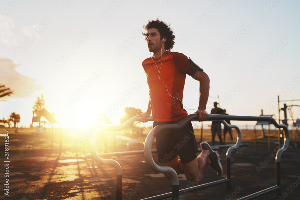 Wall mural Athletic young male athlete listening to music on earphones exercising on parallel bars doing push-ups in the park -young man doing dips outdoors