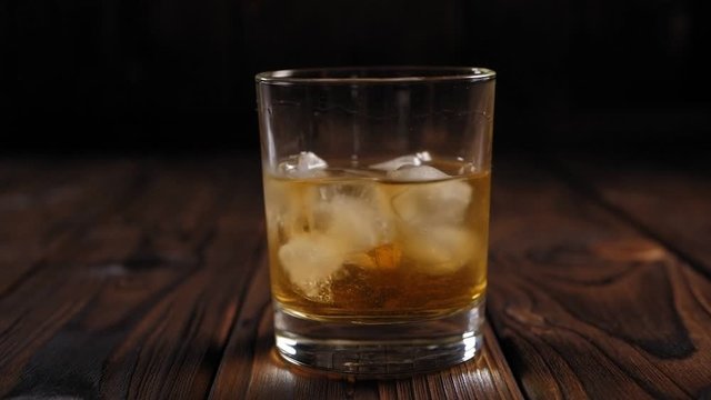Close-up of a glass of whiskey with ice, the liquid slowly rotates in the glass. A glass of chilled whiskey or cognac in a glass on a wooden background. Slow motion.