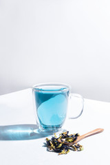 Obraz na płótnie Canvas Blue herbal Butterfly pea flower tea in a glass mug on white background. Next to the mug are dry inflorescences. Hard shadows. Minimalism.