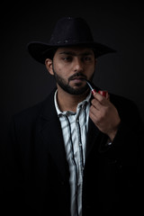 Fashion portrait of an young and handsome Indian Bengali brunette man with striped formal shirt, black suit, hat and a pipe in black copy space background. Indian lifestyle and fashion photography.