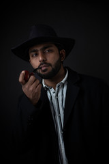 Fashion portrait of an young and handsome Indian Bengali brunette man with striped formal shirt, black suit, hat and a pipe in black copy space background. Indian lifestyle and fashion photography.