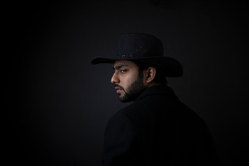 Fashion portrait of young and handsome Indian Bengali brunette man with black suit and hat turning back in black copy space background. Indian lifestyle and fashion photography.