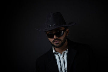 Fashion portrait of an young and handsome Indian Bengali brunette man with striped formal shirt, black suit, sunglasses and cowboy hat in black copy space background. Indian portrait