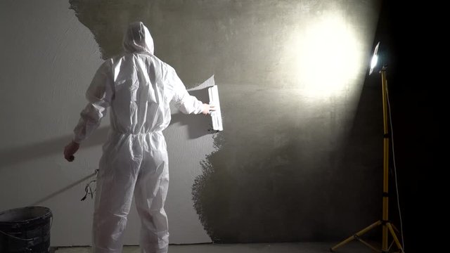 Plasterer in white working uniform plastering the wall indoors. Construction finishing works. Builder using plastering tool for finishing wall. Worker making repair in room.