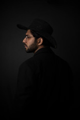 Fashion portrait of young and handsome Indian Bengali brunette man with striped formal shirt, black suit, glasses and cowboy hat in black copy space background. Indian lifestyle and fashion photograph