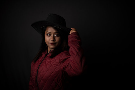 Fashion portrait of an young Indian Bengali brunette woman with maroon winter jacket and cowboy hat in black copy space background. Indian lifestyle and fashion photography.