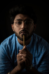 Portrait of young and handsome Indian Bengali brunette man with traditional wear and glasses holding pencil in front of his lips gesturing silence in black copy space background. Indian lifestyle