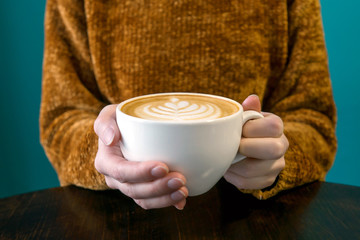 Woman in terracotta sweater headless and hands with latte. Turquoise background, brown wooden table.