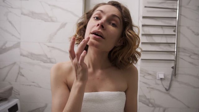 Young woman in big white towel is applying cream on the face under eye area with massage movements and looking at mirror in domestic bathroom after shower. Slow motion
