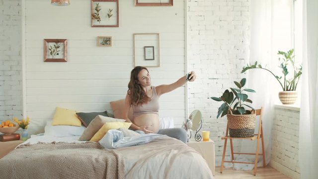 Smiling pregnant woman taking selfie photo. Expectant mother sitting in bed.