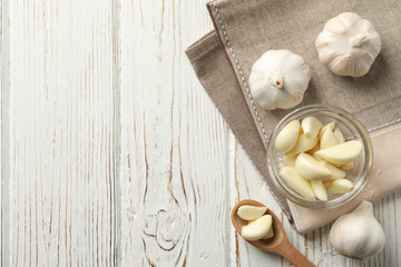 Glass bowl of fresh garlic bulbs, spoon of slices, napkin on white wooden background, space for text. Top view