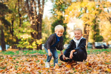 Cute two brothers playing with autumn leaves in a park. Stylish kids having fun outdoors. Autumn fashion and lifestyle.