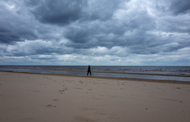 Fototapeta na wymiar A girl in a warm black jacket walks along the seashore on a sandy beach in cloudy, cool weather.