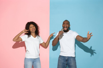 He singing, she angry. Young man and woman in casual clothes on pink, blue bicolored background. Concept of human emotions, facial expession, relations, ad. Beautiful african-american couple.