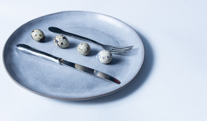 Quail eggs on gray plate between vintage fork and knife, reflecting in steel,  on white background