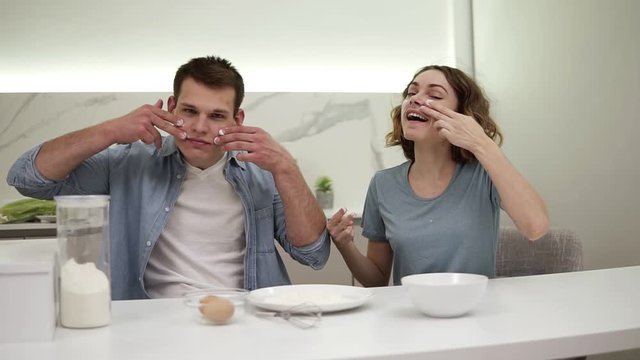 Cute positive couple smudges flour on their faces in the kitchen. Happy young couple fooling around while cooking food. Slow motion