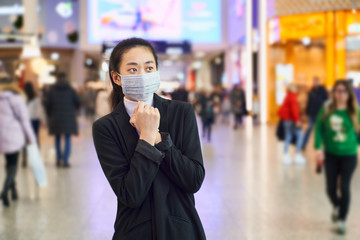 Fototapeta na wymiar Alarmed female traveler of Asian appearance wears medical mask to protect, afraid of infection, in shopping center. Deadly coronavirus in China 2019 2020 2019-nCoV. Free space for text or advertising.