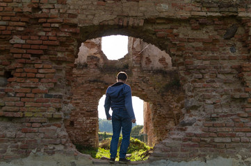 Fototapeta na wymiar A young man in blue clothes stands on the ruins of an old Golshany castle, turning his back, framed by a round hole in the castle wall