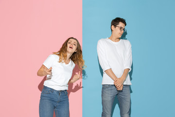 Fototapeta premium She's dancing, he's calm. Young and happy man and woman in casual clothes on pink, blue bicolored background. Concept of human emotions, facial expession, relations, ad. Beautiful caucasian couple.