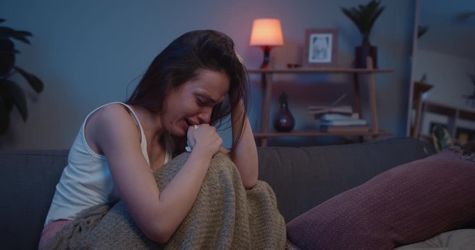 Lonely sad girl sitting on sofa in fetal position and crying. Depressed young woman in tears covered with blanket broke up with boyfriend . Concept of loneliness and relationship.