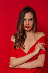 Confident pretty woman with wavy hair looking at camera wearing red dress with red lips over red background. Stylish woman's portrait