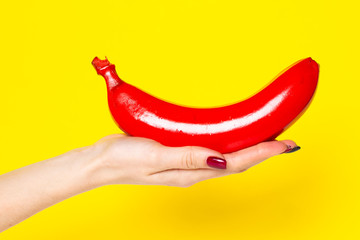 Photo of a banana painted red on a female palm. Banana in hand on a yellow background with place for text.